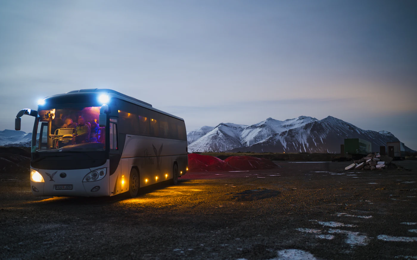 Coach bus with its lights on parked in front of snowy mountains at dusk