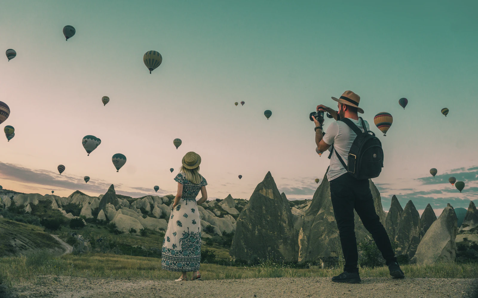 Two travelers photographing hot-air balloons over a rocky valley at sunrise
